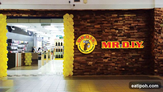 The exterior of a MR. D.I.Y. store is visible, decorated with yellow balloons on either side of the entrance. The store facade features a brick-patterned wall with the MR. D.I.Y. logo prominently displayed. Through the open entrance, shelves stocked with various products can be seen inside the store.