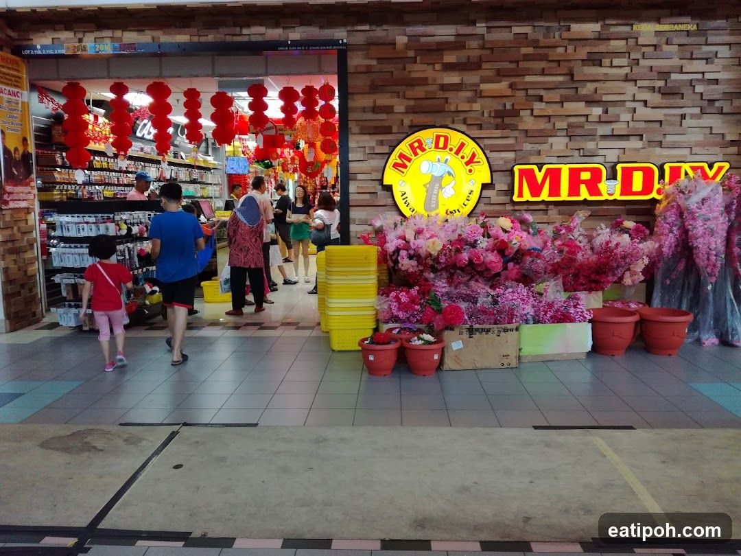 The exterior of a MR. DIY store is shown, decorated with red lanterns and artificial flowers. Several people are browsing inside the store, which is stocked with various goods. The store's signage is prominently displayed on the brick facade.