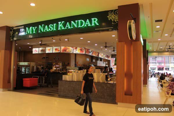 A brightly lit storefront with a large green neon sign reading 'MY NASI KANDAR' above the entrance. The sign also includes 'RESTORAN 23 JAM' to its left. A wooden pillar with a clock is on the right side of the entrance. Inside, food preparation counters and staff are visible, along with illuminated menu boards displaying images of dishes. A pedestrian walks past the entrance carrying a bag.