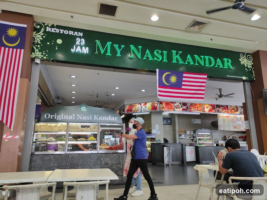 A storefront with a green neon sign reading 'MY NASI KANDAR' above the entrance. The sign also includes 'RESTORAN 23 JAM'. Two Malaysian flags are displayed on either side of the entrance. Inside, there is an open counter area with food displays under glass and a menu board mounted on the wall. Customers are seen inside and outside the restaurant.