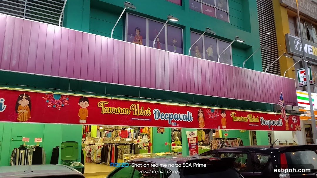 Exterior view of a multi-level retail building with green and pink facade. A large red banner spans the storefront advertising 'Tawaran Hebat Deepavali' in white and blue lettering, flanked by cartoon characters. Above, windows display mannequins wearing traditional attire. Below, clothing racks are visible inside the shop entrance. Parked cars are in foreground. Signage includes a 7-Eleven logo on a pole to the right.