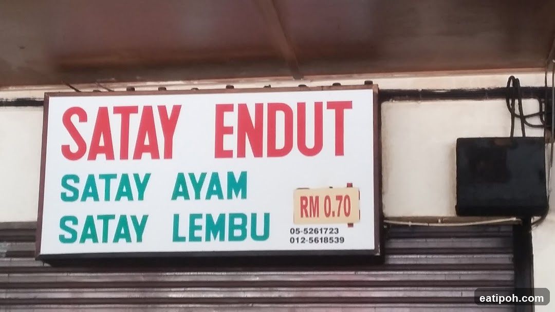 A white rectangular sign with red and green lettering mounted above a closed metal roller shutter door. The sign reads 'SATAY ENDUT' in large red letters at the top, followed by 'SATAY AYAM' and 'SATAY LEMBU' in green below it. To the right is a small yellow price tag showing 'RM 0.70'. Below the main text are two phone numbers: '05-5261723' and '012-5618539'. The sign is affixed to a light-colored wall under a dark brown ceiling or awning.