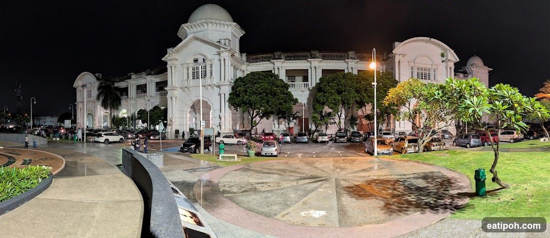 Night view of a grand white building with a dome and arched entrances, surrounded by trees and a parking lot filled with cars.