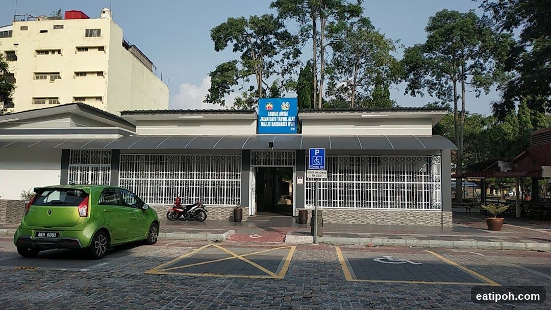 The exterior of a public toilet facility with a blue sign above the entrance. The sign reads 'TANDAS AWAM JALAN DATO TAHWIL AZAR MAJLIS BANDARAYA IPOH 2010'. There are parking spaces, including one for disabled individuals, in front of the building. A green car and a motorcycle are parked nearby.