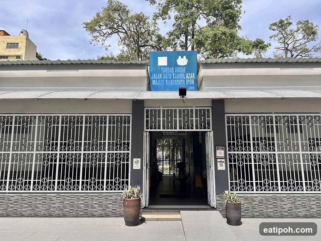 The exterior of a public restroom facility with a gray facade, white barred windows, and a blue sign above the entrance. The sign reads "TANDAS AWAM JALAN DATO TAHWIL AZAR MAJLIS BANDARAYA IPOH 2018".