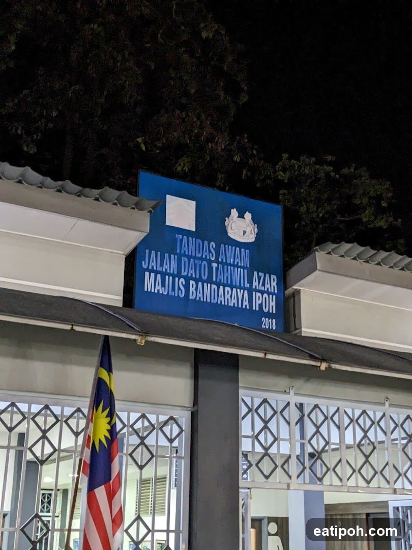 A blue sign with white text reads "TANDAS AWAM JALAN DATO TAHWIL AZAR MAJLIS BANDARAYA IPOH 2018". The Malaysian flag is displayed on a pole in front of a building with a patterned metal gate.