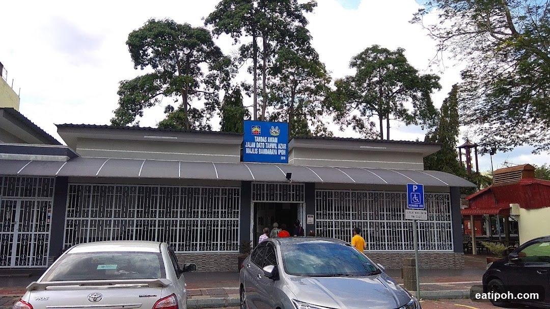 The exterior of a government building with a prominent blue sign above the entrance. The sign displays text in Malay and a coat of arms. The building has large windows covered with decorative metal grilles. Several cars are parked in front, and a few people are visible entering the building. A parking sign for disabled individuals is also present.