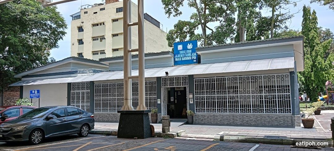 A blue sign with white text and symbols is mounted above the entrance of a building. The sign reads "TANDAS AWAM JALAN DATO TAHWIL AZAR MAJLIS BANDARAYA IPOH". The building has a grey facade with white decorative bars on the windows and a grey roof. A dark grey car is parked in front of the building.