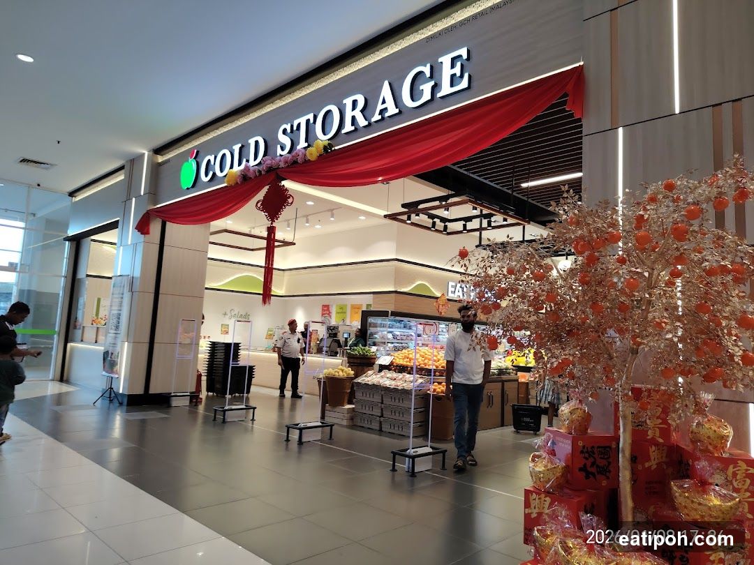 The exterior entrance of a Cold Storage supermarket, decorated with red drapes and festive ornaments, possibly for a holiday celebration. An artificial tree with orange fruits and gift boxes are displayed near the entrance.