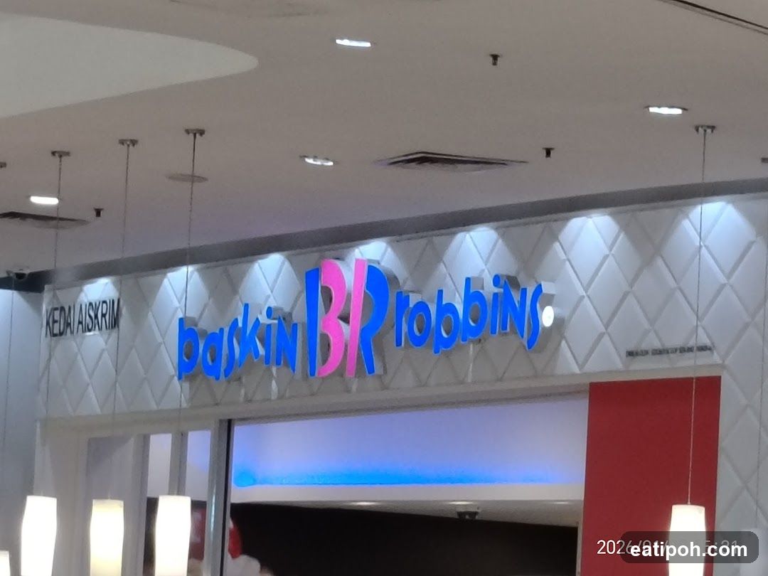 The exterior of a Baskin Robbins ice cream shop. The prominent blue and pink "Baskin Robbins" logo is displayed above the entrance. To the left of the logo, "KEDAI AISKRIM" is visible, which translates to "Ice Cream Shop" in Malay. The storefront features a white, diamond-patterned facade with integrated lighting.