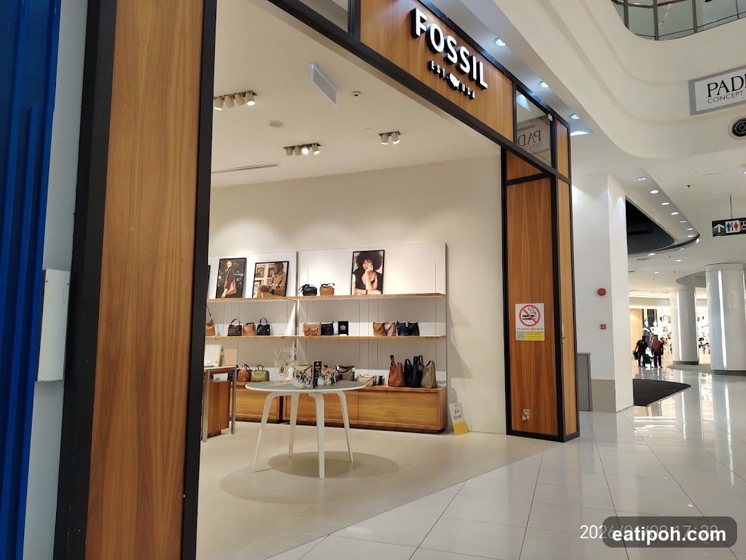 The exterior of a Fossil store with its prominent signage above the entrance. The store displays handbags and accessories on shelves and a round table. The interior is visible through the open entrance, showing a well-lit retail space.