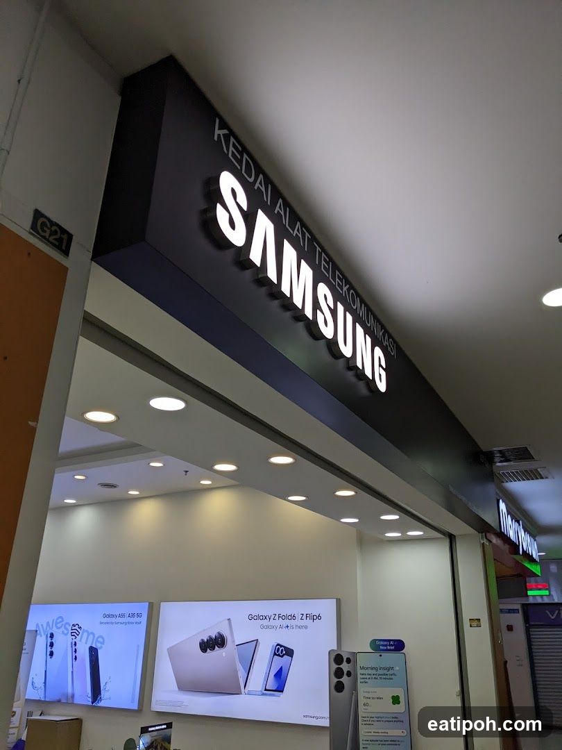 The exterior of a Samsung telecommunications store. A large illuminated sign above the entrance reads "SAMSUNG KEDAI ALAT TELEKOMUNIKASI". Inside, displays show Samsung Galaxy phones, including the Galaxy A55/A35 5G and Galaxy Z Fold6/Z Flip6.