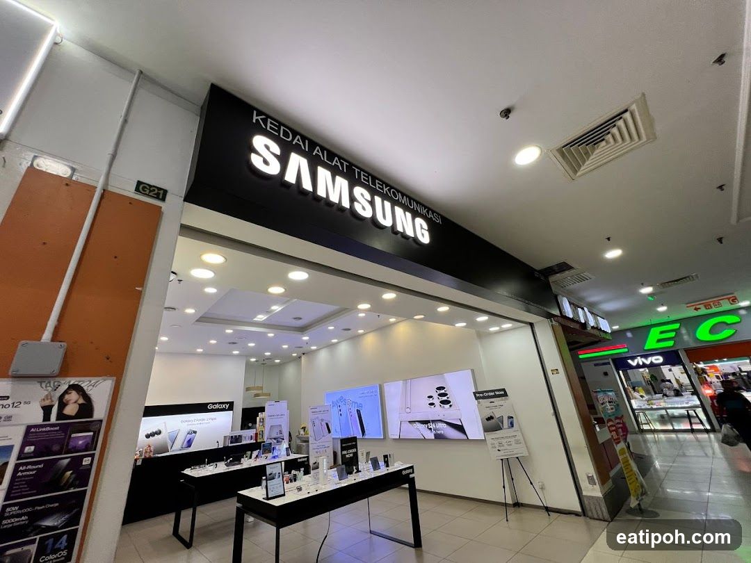 The exterior of a Samsung electronics store. A large illuminated sign above the entrance reads "KEDAI ALAT TELEKOMUNIKASI SAMSUNG". Inside, display shelves showcase various Samsung mobile phones and accessories. The store is located in a mall, with other businesses visible next door.