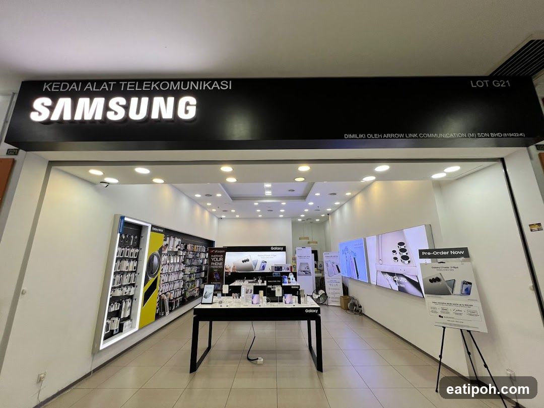The exterior of a Samsung electronics store, with the Samsung logo prominently displayed above the entrance. Inside, various Samsung products and accessories are displayed on shelves and tables. Digital screens showcase product images and promotions.