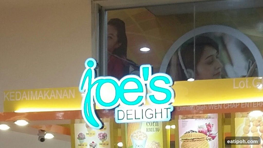 A bright, illuminated sign for "Joe's Delight" is prominently displayed above a food stall. The sign features the name in a stylized, turquoise font. Below the main sign, the word "DELIGHT" is also illuminated. A yellow banner with the word "KEDAI MAKANAN" is visible to the left of the main sign. The stall displays images of food items, including ice cream, waffles, and what appears to be popcorn. The overall impression is of a cheerful and inviting dessert or snack shop.