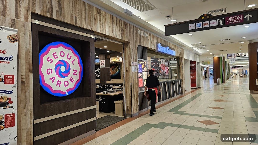 The exterior of a Seoul Garden restaurant in a mall. A large circular sign with the restaurant's name is visible on the left. The entrance to the restaurant shows tables and seating inside. A staff member in uniform is walking past the counter area. Signs and menus are displayed on the wall outside the restaurant.