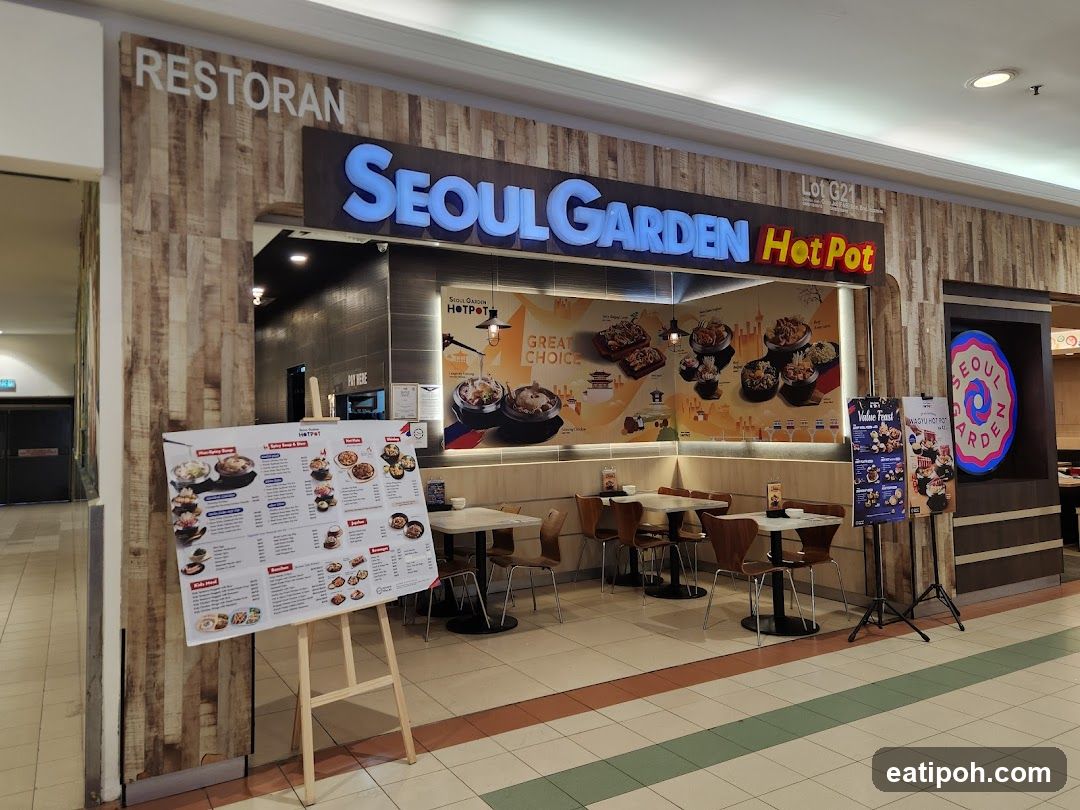 The exterior of a Seoul Garden Hot Pot restaurant. A large sign above the entrance reads "SEOUL GARDEN Hot Pot". In front of the restaurant, a menu is displayed on an easel. Inside, tables and chairs are visible, along with a mural depicting food.