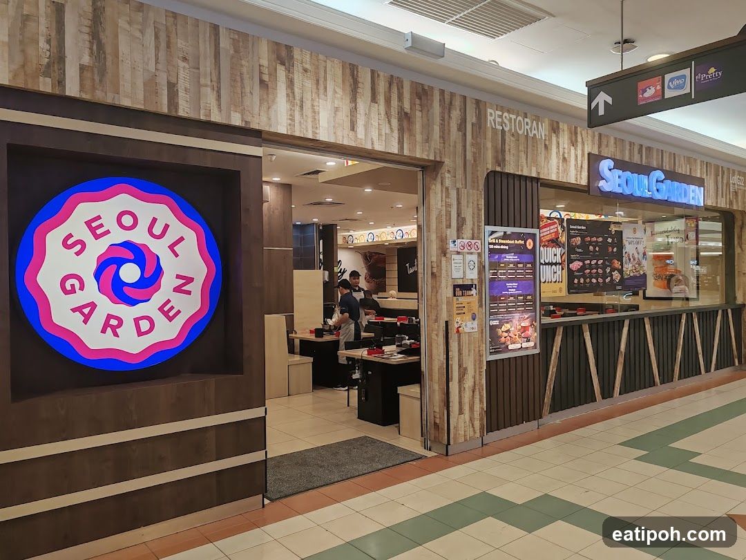 The exterior of a Seoul Garden restaurant with a large circular logo on the left and the restaurant name in illuminated blue letters above the counter. The interior is visible through the glass, showing tables and staff. There are also promotional posters and menus displayed.
