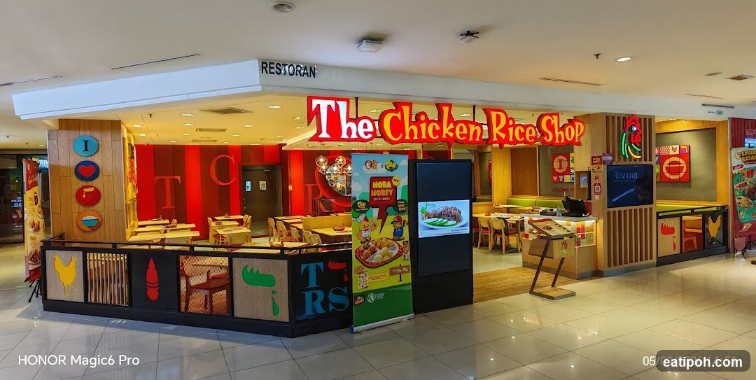 The exterior of a restaurant called "The Chicken Rice Shop". The storefront features a prominent red and yellow sign with the restaurant's name. Inside, tables and chairs are visible, along with decorative elements like colorful circles with letters and icons. A digital display screen and a promotional banner are placed near the entrance.