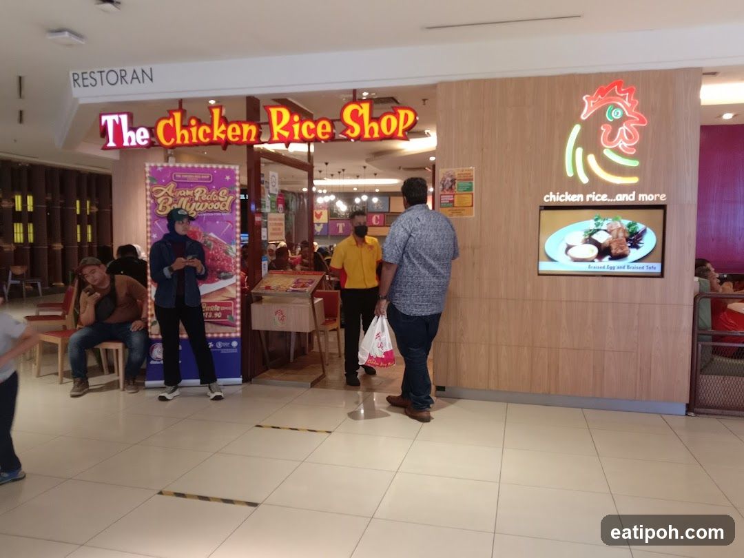 The exterior of a restaurant called "The Chicken Rice Shop". A sign above the entrance reads "RESTORAN The Chicken Rice Shop". To the right, a neon chicken logo is displayed above a screen showing a dish called "Braised Egg and Braised Tofu".