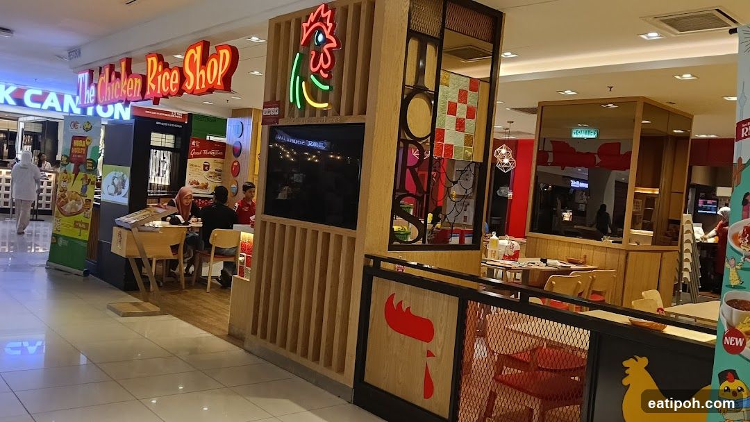The exterior of a restaurant called "The Chicken Rice Shop" is visible. The shop has a prominent red and yellow sign with a neon chicken logo. Inside, tables and chairs are set up for dining. There are also promotional posters and displays visible.