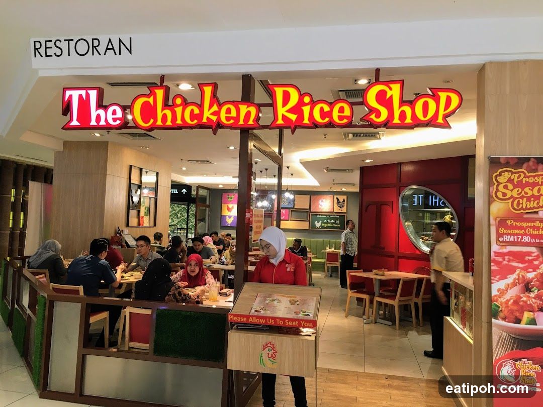The exterior of a restaurant called "The Chicken Rice Shop". The restaurant has a red and yellow sign with the name of the shop. Inside, customers are seated at tables and eating. There is also a promotional poster on the right side of the image.