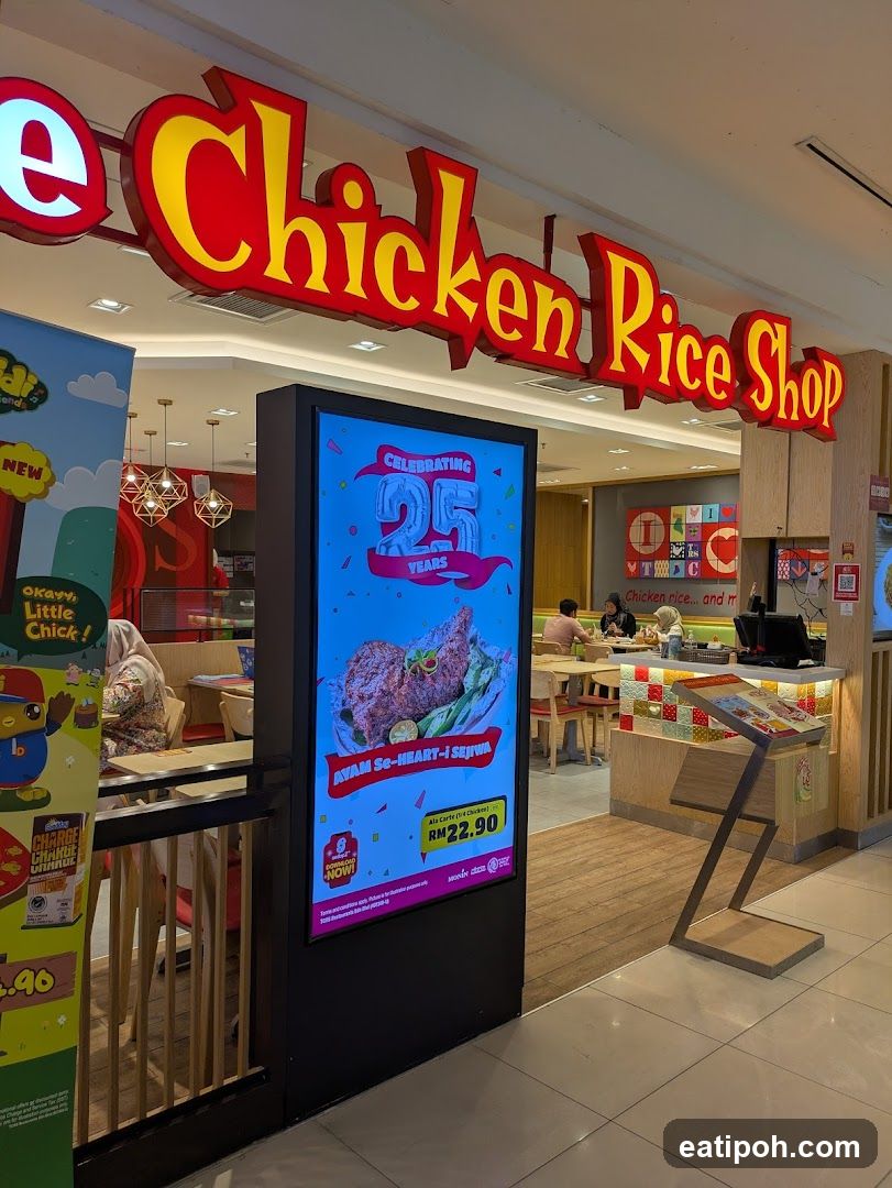 The exterior of The Chicken Rice Shop with a large red and yellow sign above the entrance. A digital display shows a promotional image of chicken and text that reads "CELEBRATING 25 YEARS" and "AVAM Se-HEART-I SEJIWA RM22.90". Inside, diners are seated at tables.