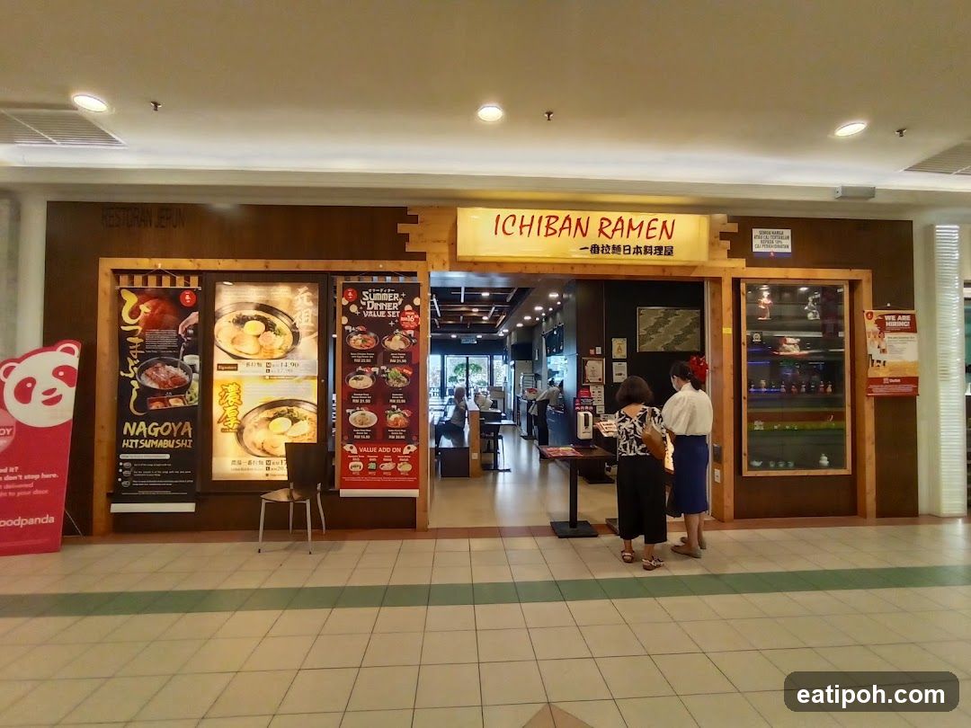 The exterior of a ramen restaurant with signage above the entrance. Posters displaying food items and promotions are visible on the left and right sides of the entrance. Two people are standing near the entrance.