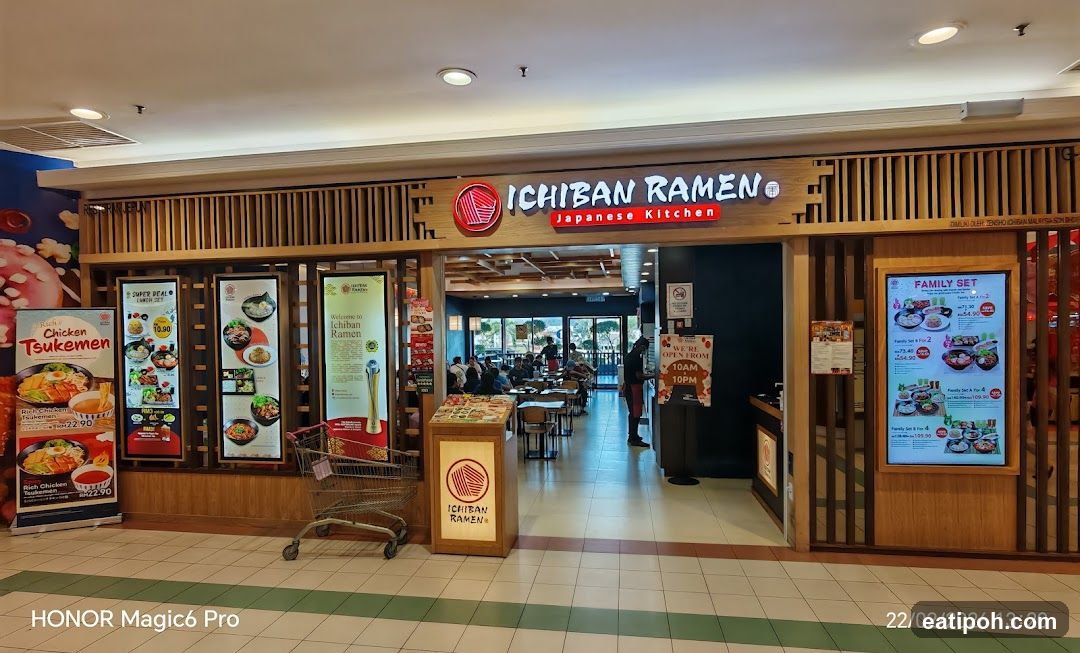 The exterior of Ichiban Ramen, a Japanese kitchen, with its prominent signage above the entrance. Various promotional displays and menus are visible outside the restaurant, showcasing dishes like Tsukemen and family sets. The interior of the restaurant is visible through the open entrance, with patrons seated at tables.