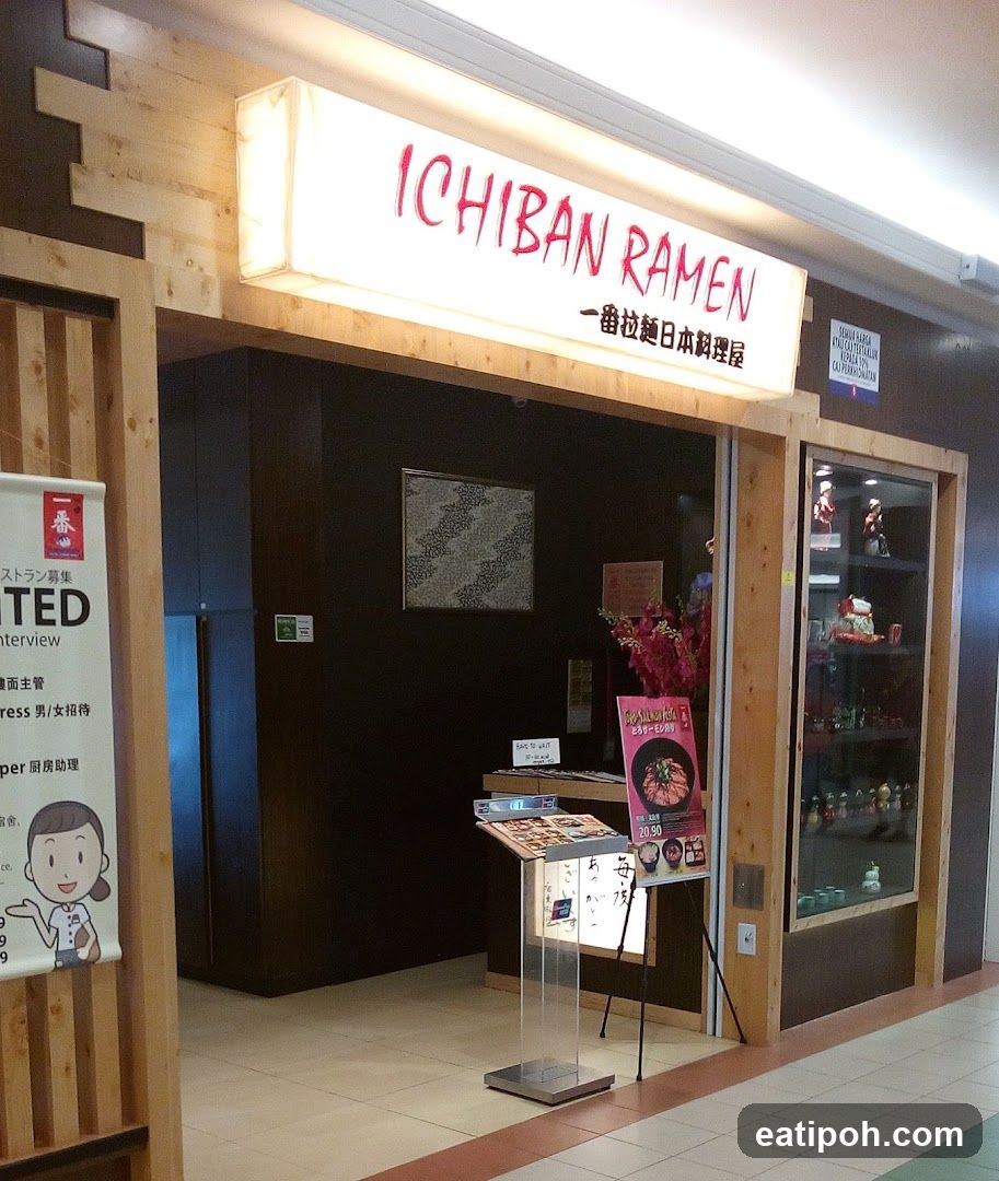 The exterior of a ramen restaurant with a prominent illuminated sign that reads "ICHIBAN RAMEN" in red lettering, with Japanese characters below. The entrance is framed by wooden accents. To the left, a banner with a cartoon chef and Japanese text is visible. Inside, a display stand with a menu and a promotional poster featuring a ramen dish are present. A glass display cabinet with figurines is to the right.