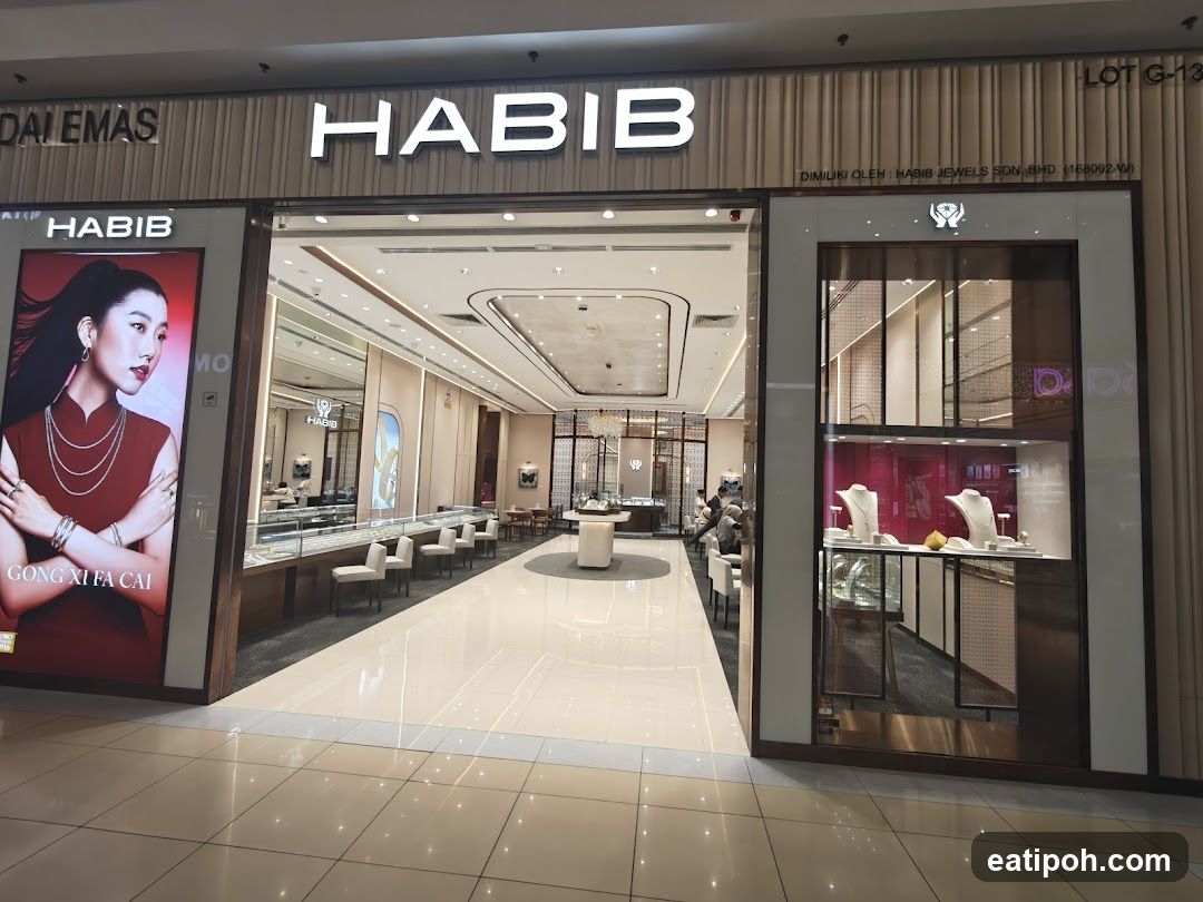 The exterior of a Habib Jewels store, a jewelry shop. The storefront features a large illuminated sign with the name "HABIB" prominently displayed. To the left, a large advertisement poster shows a woman wearing jewelry with the text "GONG XI FA CAI". The entrance is open, revealing a well-lit interior with display cases filled with jewelry, seating areas, and more display shelves. To the right, a display window showcases jewelry on mannequins. The overall impression is one of luxury and elegance.