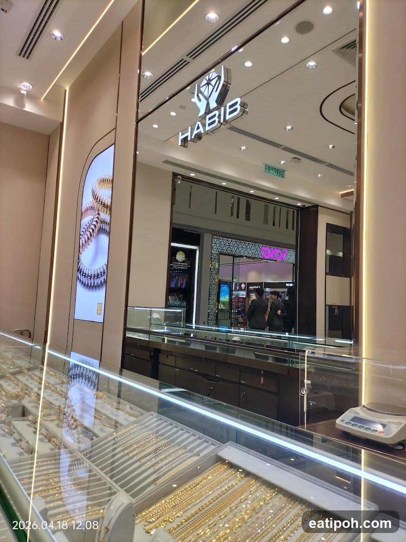 The image shows the interior of a jewelry store with display cases filled with gold necklaces and bracelets. The store's name, "HABIB," is prominently displayed above the entrance, along with a logo featuring hands holding a diamond. A digital screen on the left displays an image of a gold bracelet. In the background, customers are seen browsing.