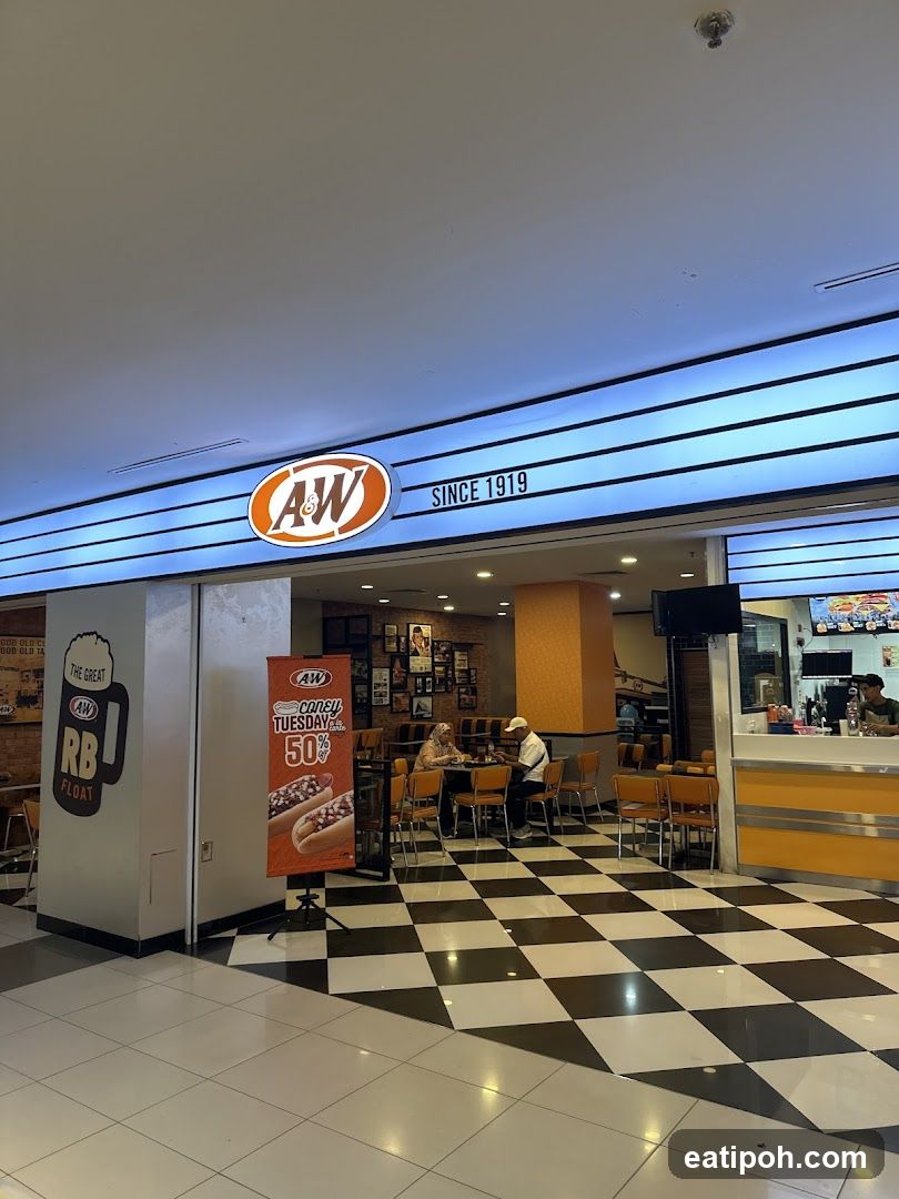 The exterior of an A&W restaurant with its prominent orange and white logo and "SINCE 1919" text. The entrance features a checkered black and white floor. Inside, patrons are seated at tables. A promotional banner for "Coney Tuesday" with a 50% discount is displayed near the entrance.