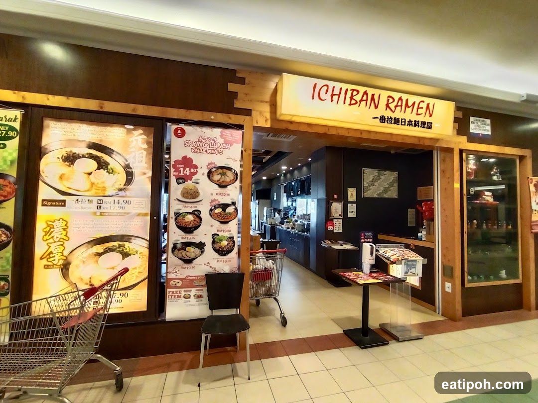 The exterior of a ramen restaurant with a prominent illuminated sign that reads "ICHIBAN RAMEN" and Japanese characters below. To the left, large posters display images of ramen bowls and prices. A "Spring Lunch Value Meals" poster is also visible. The entrance is open, revealing a glimpse of the interior seating and counter area.