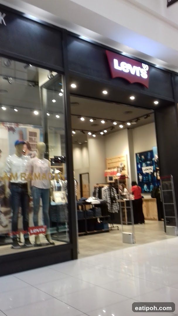 The exterior of a Levi's store. The iconic red Levi's logo is visible above the entrance. Mannequins dressed in jeans and shirts are displayed in the storefront window. Inside, clothing racks and a checkout counter are visible.