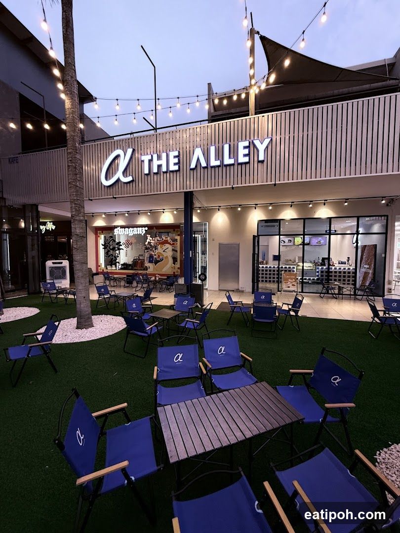 A storefront with the name "THE ALLEY" in large illuminated letters above a row of windows. The area in front of the store is covered in artificial grass and furnished with blue chairs and wooden tables. String lights are hung overhead.