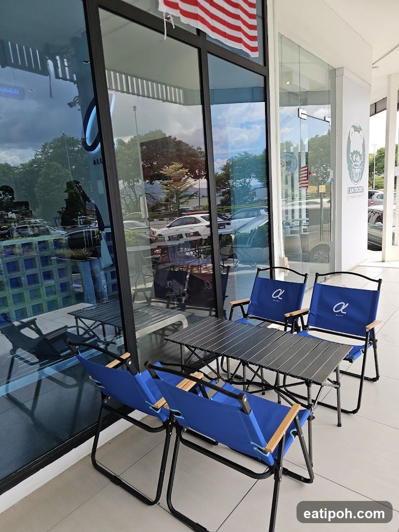 A storefront with large glass windows and an outdoor seating area. The seating area includes several blue folding chairs and a portable table, all branded with a logo. Reflections in the glass show trees, cars, and the sky. A sign with a cartoon character and text is visible on the right side of the storefront.