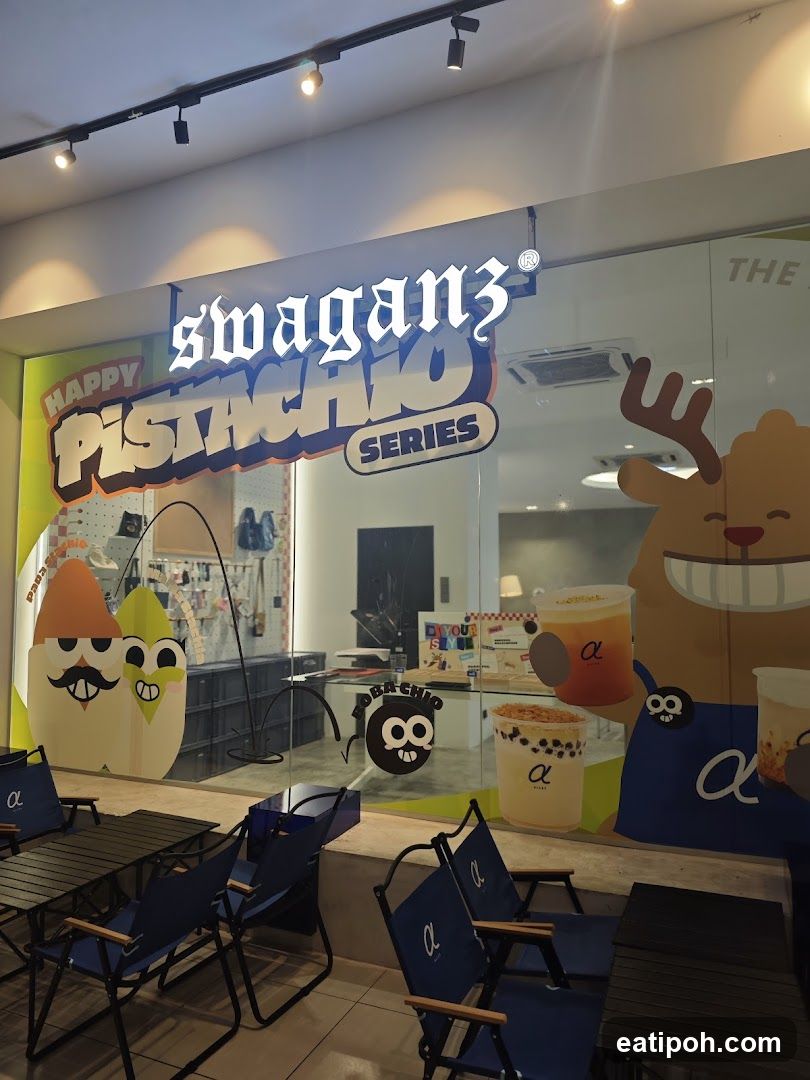 The storefront of a shop called Swaganz, featuring a large illuminated sign with the text "swaganz" and "HAPPY PISTACHIO SERIES". The glass facade displays cartoon characters, drinks, and the shop's logo. Outdoor seating with blue chairs and tables is visible in front of the shop.