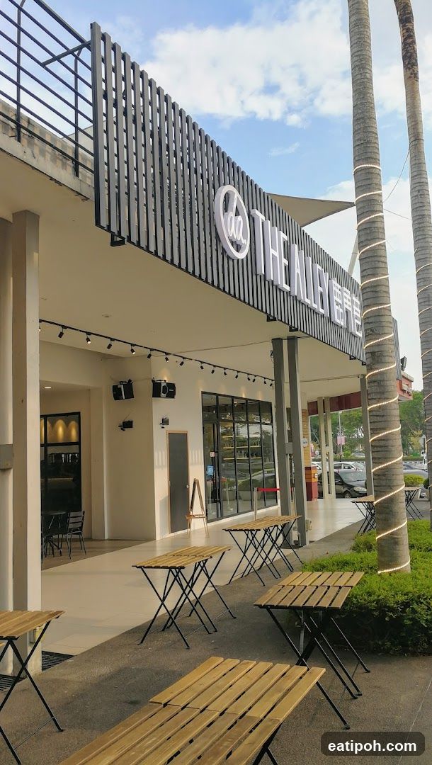 The exterior of a shop with the name "THE ALLEY" prominently displayed on a sign above the entrance. The shop has large glass windows and an outdoor seating area with several wooden tables and chairs. Palm trees decorated with string lights are visible to the right of the storefront.