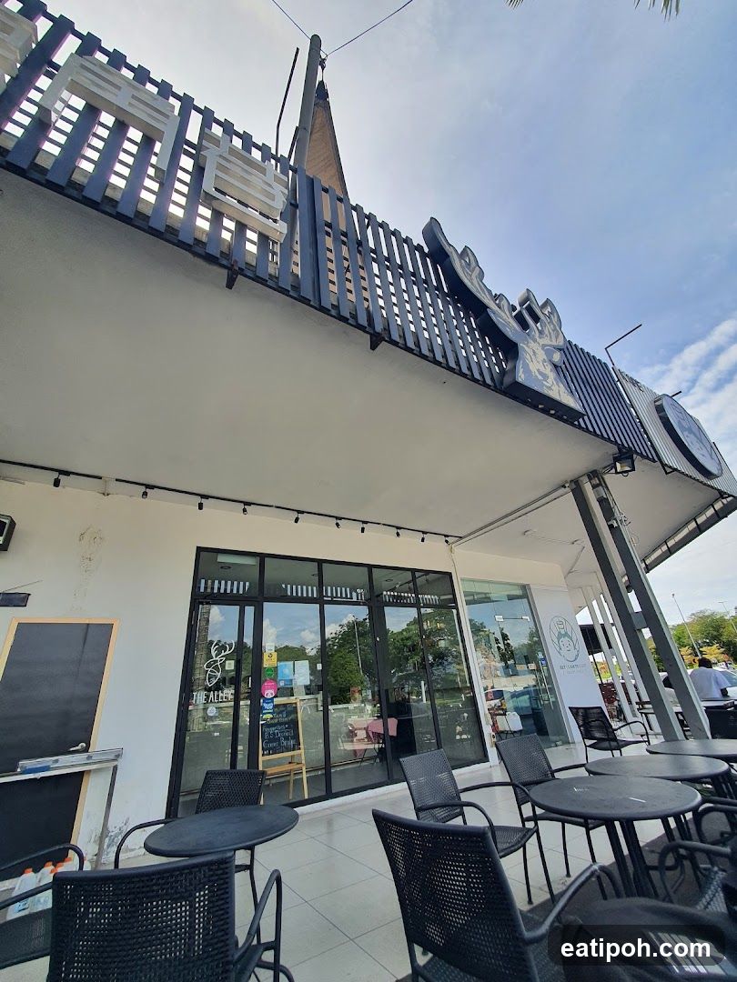 The exterior of a modern cafe with large glass windows and doors. Outdoor seating with black tables and chairs is visible on the patio. A prominent sign with the name "THE ALLEY" is displayed on the glass facade. Above the cafe, a large, stylized sign with Asian characters is mounted on the building's overhang.