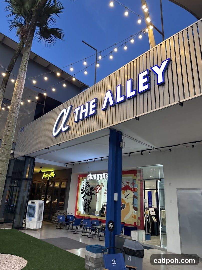 The exterior of a store with a prominent sign that reads "THE ALLEY" in large blue letters. The storefront features a modern design with vertical metal siding and string lights overhead. To the left, another establishment with a sign reading "Pretty Me" is visible. In front of the stores, there are several blue chairs and tables set up, suggesting an outdoor seating area. A large air cooler unit is placed to the left of the entrance. The overall scene is set against a twilight sky.