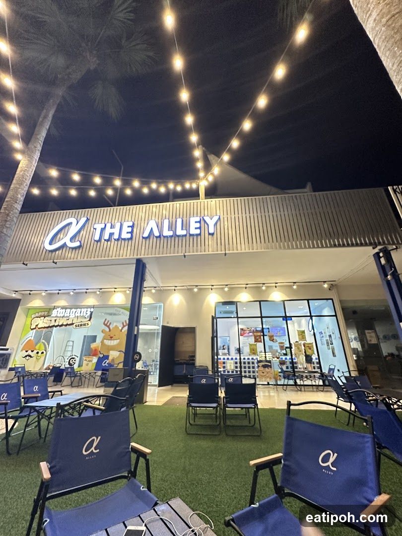 The exterior of a shop called The Alley at night. String lights are hung above the entrance. There are outdoor seating areas with blue chairs and tables on artificial grass. The shop has large glass windows and a sign above the entrance that reads "THE ALLEY" with a logo.