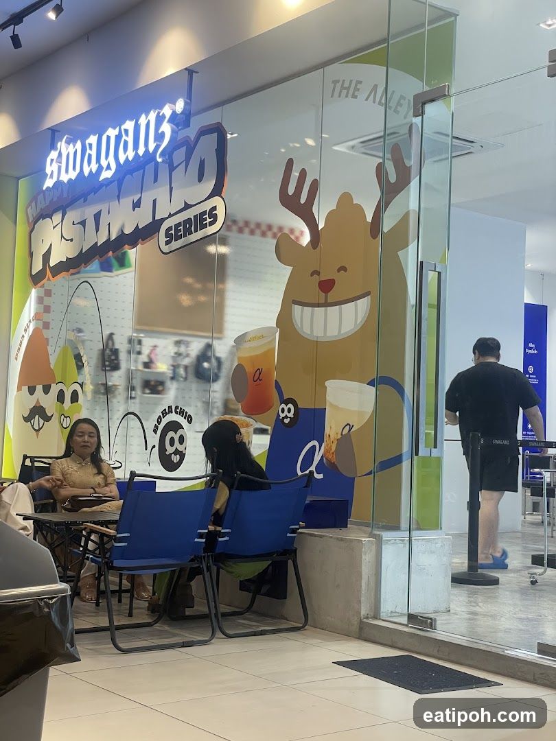 The exterior of a shop with large glass windows displaying colorful graphics and signage. The shop's name, "swaganz HAPPY PISTACHIO SERIES," is prominently displayed on the window. Inside, people are seated at tables, and a cartoon character with antlers is visible. A man in a black shirt and shorts is walking out of the shop.